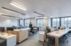 Ein lichtdurchflutetes, modernes Büro, in dem eine diverse Führungskraft mit einem konzentrierten, freundlichen Lächeln vor mehreren Bildschirmen sitzt, während im Hintergrund teamorientierte Gespräche und digitale Datenvisualisierungen in warmem Tageslicht eine harmonische Atmosphäre von innovativer Zusammenarbeit zwischen Mensch und KI vermitteln.