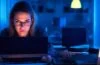 In einem hell erleuchteten, modernen Büro sitzt eine junge Frau konzentriert vor einem Laptop, die Hände über der Tastatur, während natürliches Tageslicht ihr Gesicht sanft wärmt und im Hintergrund unscharf Bücherregale voller Fachliteratur sowie ein Glas Wasser auf dem Tisch für eine authentische Arbeitsatmosphäre sorgen.