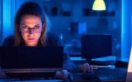In einem hell erleuchteten, modernen Büro sitzt eine junge Frau konzentriert vor einem Laptop, die Hände über der Tastatur, während natürliches Tageslicht ihr Gesicht sanft wärmt und im Hintergrund unscharf Bücherregale voller Fachliteratur sowie ein Glas Wasser auf dem Tisch für eine authentische Arbeitsatmosphäre sorgen.