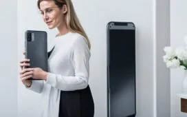 Eine einladende, helle Szene mit einer Person, die entspannt modern wirkende Smartphones in der Hand hält, umgeben von warmem Tageslicht an einem minimalistischen Holzschreibtisch, während im Hintergrund verschwommen helle Fenster und dezente Pflanzen eine freundliche, vertrauensvolle Atmosphäre für digitale Kommunikation und Datenschutz schaffen.