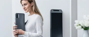 Eine einladende, helle Szene mit einer Person, die entspannt modern wirkende Smartphones in der Hand hält, umgeben von warmem Tageslicht an einem minimalistischen Holzschreibtisch, während im Hintergrund verschwommen helle Fenster und dezente Pflanzen eine freundliche, vertrauensvolle Atmosphäre für digitale Kommunikation und Datenschutz schaffen.