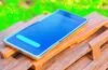 Ein helle, sonnendurchflutete Nahaufnahme eines modernen Smartphones auf einem Holztisch, umgeben von entspannt lächelnden Menschen in heller Kleidung, die gemeinsam konzentriert ihre Geräte sichern – ein warmes, freundliches Setting voller natürlicher Lichtreflexe, das Vertrauen und digitale Sicherheit harmonisch vermittelt.