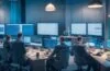 Ein hell erleuchteter, moderner Bankarbeitsplatz mit einem konzentrierten Team aus Mitarbeitenden, die gemeinsam an Bildschirmen mit komplexen Daten und Sicherheitsanalysen arbeiten, in warmer Tageslichtatmosphäre, die Vertrauen und technische Kompetenz ausstrahlt.
