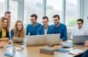 Ein warmes, sonnendurchflutetes Büro mit einem fokussierten, vielfältigen Team aus jungen Erwachsenen und älteren Menschen, die gemeinsam an Laptops arbeiten und sich mit freundlichen Gesten über innovative KI-Technologien austauschen – ein lebendiges Sinnbild für digitale Teilhabe und zukunftsweisende Zusammenarbeit in Großbritannien.