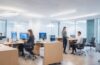 Helles, modernes Büro mit warmem Tageslicht, in dem ein selbstbewusst lächelnder Manager während eines Gespräches mit seinem Team strategisch auf digitale Finanzdaten auf einem Laptop blickt – ein Bild voller positiver Energie, das Aufbruchsstimmung und innovative Führung im Kontext eines bevorstehenden Börsengangs ausstrahlt.