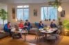 Eine helle, einladende Szene in einem modernen Wohnzimmer, in dem eine vielfältige Gruppe junger Menschen entspannt mit Laptops und Smartphones um einen gemütlichen Tisch sitzt, harmonisch von warmem Tageslicht durchflutet, das die digitale Vernetzung und den globalen Preiskampf der Streaming-Dienste spürbar macht.