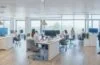 Ein modernes, lichtdurchflutetes Büro mit freundlichen Menschen verschiedener Altersgruppen, die entspannt und konzentriert mithilfe von Smartphones und Laptops eine sichere Zwei-Faktor-Authentifizierung durchführen, während warmes Tageslicht das helle, aufgeräumte Ambiente durchflutet und eine Atmosphäre von Vertrauen und digitaler Sicherheit vermittelt.