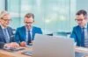 Helle, freundliche Büroszene mit mehreren konzentrierten Fachleuten in moderner Geschäftskleidung, die lebhaft an digitalen Geräten arbeiten und gemeinsam Strategien zur IT-Sicherheit und Compliance im Finanzsektor besprechen, eingefangen in warmem, natürlichem Licht mit realistischen Details und einem klaren, einladenden Ambiente.