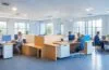 Ein lichtdurchflutetes, modernes Büro mit mehreren engagierten Mitarbeitenden verschiedenen Alters und Geschlechts, die gemeinsam an Laptops und Tablets mit digital unterstützten Arbeitsprozessen konzentriert und freundlich lächelnd eine neue KI-Anwendung ausprobieren, während warmes Tageslicht durch große Fenster fällt und eine produktive, zukunftsorientierte Atmosphäre schafft.