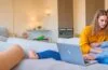 Eine warme, helle Szene eines konzentrierten jungen Erwachsenen an einem sonnendurchfluteten Schreibtisch, der engagiert auf einen Laptopbildschirm blickt, umgeben von moderner Alltagstechnik und subtilen Sicherheitshinweisen, die ein Gefühl von Achtsamkeit und digitaler Vorsicht vermitteln.