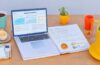 Ein heller, freundlich illuminierter moderner Arbeitsplatz mit einem aufgeschlagenen Laptop, auf dessen Bildschirm Diagramme und Webinhalte zu sehen sind, umgeben von Notizbüchern und einer tasse Kaffee, während weiches Tageslicht durch ein großes Fenster fällt und eine Atmosphäre von Innovation und digitaler Vernetzung vermittelt.