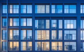 In einem lichtdurchfluteten, modernen Bürogebäude tauschen sich diverse Fachleute lebhaft über digitale Infrastrukturprojekte aus, während große Fenster warme Sonneneinstrahlung hereinlassen und im Hintergrund schemenhaft Serverracks und Netzwerkkomponenten zu erkennen sind – eine harmonische Verbindung von Mensch, Technologie und Zukunftsvision.