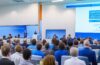 Ein heller, inspirierender Konferenzraum mit modernen, transparenten Bildschirmen, auf denen schemenhafte, digitale Avatare von Wissenschaftlern in lebhaftem Austausch stehen, umgeben von natürlichem Tageslicht, das die freundliche und zukunftsweisende Atmosphäre eines virtuellen KI-Gipfels warm und realistisch einfängt.