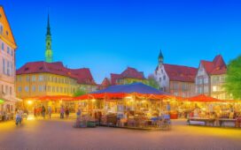 Ein sonnendurchfluteter, belebter Marktplatz in einer niedersächsischen Stadt, auf dem moderne Sicherheitskameras diskret an Laternenmasten montiert sind, während freundliche Passanten in entspannter Atmosphäre ihrem Alltag nachgehen und die warme, natürliche Lichtstimmung Vertrauen und Dialogbereitschaft zwischen Technologie und Gesellschaft symbolisiert.