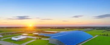 Ein strahlendes Foto bei warmem Tageslicht zeigt den modernen Maincubes-Datacenter-Campus in Nauen mit klaren Linien und nachhaltigen Solarpanels, umgeben von weiten grünen Flächen, während im Hintergrund die Silhouette Berlins in sanften Morgenfarben leuchtet und ein Gefühl von Zukunft, Klarheit und technologischer Innovation vermittelt.