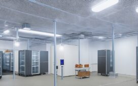 Ein warm ausgeleuchteter, moderner Serverraum mit klar strukturierten Elektroinstallationen und leuchtenden Kontrolllampen, in dem technisches Fachpersonal aufmerksam und konzentriert die Sicherheitsstandards der VDE-Normen überprüft, eingebettet in eine harmonische Atmosphäre aus natürlichem Tageslicht und funktionaler Eleganz.