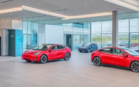 Ein sonnendurchfluteter, moderner Showroom mit zwei eleganten Elektroautos – ein silbernes Tesla Model Y und ein leuchtend rotes BYD-Modell – die Seite an Seite im Fokus stehen, umgeben von warmen Holztönen und lebendigem Grün, strahlen technologische Innovation und freundliche Zukunftsvision aus.