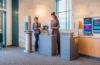 Ein einladender Hotel-Empfangsbereich bei Tageslicht, in dem eine freundliche Rezeptionistin mit einem gastfreundlichen Lächeln an einem modernen Self-Check-in-Terminal assistiert, während dezente Kameras und Sensoren im Hintergrund die Balance zwischen technologischer Überwachung und warmem, persönlichem Service widerspiegeln.