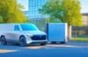 Ein strahlend sonnendurchfluteter europäischer Stadthintergrund, vor dem ein moderner, vollelektrischer US-Pickup mit markanter, futuristischer Linienführung wie der Tesla Cybertruck freundlich und kraftvoll in Szene gesetzt ist, während Passanten neugierig und entspannt vorbeigehen – ein lebendiges Bild von Innovation, Wandel und urbaner Mobilität im Aufbruch.