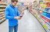 In einem sonnendurchfluteten, modernen Supermarkt zeigt eine freundliche junge Kundin mit Smartphone in der Hand entspannt den digitalen Zwei-Faktor-Authentifizierungs-Code auf dem Display, während freundliches Personal im Hintergrund mit natürlichem Tageslicht die Szene warm und einladend beleuchtet.