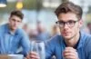 Ein sonnendurchflutetes Porträt eines jungen, sympathischen Mannes mit moderner, schlanker Smart-Brille, der entspannt in einem urbanen Café sitzt und vertrauensvoll in die Ferne blickt, umgeben von warmen Holztönen und natürlichem Tageslicht, das seine offene, zukunftsorientierte Ausstrahlung unterstreicht.