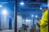 Ein hell erleuchteter, moderner Arbeitsplatz mit einem Smartphone im Vordergrund, auf dem sanftes Tageslicht fällt, während eine entspannte Person im Hintergrund konzentriert arbeitet – die Atmosphäre strahlt Vertrautheit, technische Präsenz und vorsichtige Achtsamkeit aus.