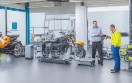 Ein strahlend sonnendurchflutetes Labor mit modernen Elektrofahrzeugen, deren offene Motorhauben Einblicke in glänzende, innovative Batteriemodule bieten, während vorsichtig forschende Hände und natürliche Grüntöne im Hintergrund die harmonische Verbindung von Technik, Nachhaltigkeit und Zukunftsinnovation verkörpern.