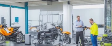 Ein strahlend sonnendurchflutetes Labor mit modernen Elektrofahrzeugen, deren offene Motorhauben Einblicke in glänzende, innovative Batteriemodule bieten, während vorsichtig forschende Hände und natürliche Grüntöne im Hintergrund die harmonische Verbindung von Technik, Nachhaltigkeit und Zukunftsinnovation verkörpern.