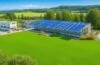 Ein strahlend sonnendurchfluteter, moderner Rechenzentrums-Campus inmitten grüner Landschaft, umgeben von nachhaltigen Solarpaneelen und Windrädern, wobei gut gekleidete Technik-Experten in angeregtem Austausch vor den großen Glasfassaden stehen und so die Zukunft Europas digitale Infrastruktur in warmem, authentischem Licht lebendig werden lassen.