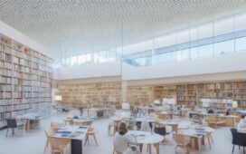 Ein sonnendurchfluteter, moderner Bibliotheksraum in Berlin mit offenen Fenstern, an hellen Holztischen sitzen lachende Menschen unterschiedlichster Altersgruppen, vertieft in digitale Geräte und Bücher, während im Hintergrund hohe Regale voller Bücher und dezente Glasfaserkabelverbindungen eine Atmosphäre von Innovation, Bildung und gemeinschaftlichem Aufbruch schaffen.