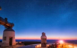 Ein warm erleuchtetes, natürlich wirkendes Editorial-Porträt eines freundlichen Astrophysikers im modernen Observatorium, der mit nachdenklichem Blick durch ein großes Panoramafenster die strahlende, sternenklare Nacht und die ferne Milchstraße betrachtet, deren funkelnde Lichter die Verbindung zwischen kosmischer Explosion und der Entstehung von Gold und Platin symbolisieren.