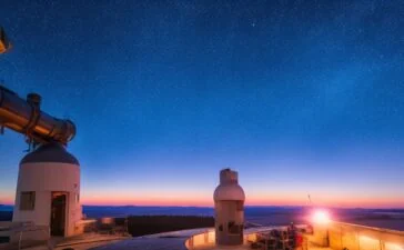 Ein warm erleuchtetes, natürlich wirkendes Editorial-Porträt eines freundlichen Astrophysikers im modernen Observatorium, der mit nachdenklichem Blick durch ein großes Panoramafenster die strahlende, sternenklare Nacht und die ferne Milchstraße betrachtet, deren funkelnde Lichter die Verbindung zwischen kosmischer Explosion und der Entstehung von Gold und Platin symbolisieren.
