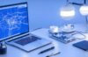 Ein heller, natürlich beleuchteter Arbeitsplatz mit einem modernen Laptop, auf dessen Bildschirm schematisch vernetzte, pulsierende Knotenpunkte eines neuronalen Netzes sichtbar sind, während im Hintergrund durch das Sonnenlicht sanft warme Holztöne und grüne Pflanzen für eine einladende, zukunftsweisende Atmosphäre sorgen.