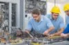 Ein heller, warm ausgeleuchteter Büroarbeitsplatz mit lächelnden IT-Experten, die konzentriert an modernen Laptops arbeiten, umgeben von dezent sichtbarer Server-Hardware und digitalen Netzwerkvisualisierungen, die eine freundliche Atmosphäre von Teamarbeit und technischer Sicherheit vermitteln.