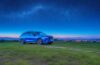 Ein strahlender E-SUV steht bei sommerlichem Tageslicht auf einer weiten Landstraße, umgeben von sanften Hügeln und blauem Himmel, während eine Familie entspannt und begeistert in das Fahrzeug einsteigt – ein Symbol für moderne Elektromobilität mit großer Reichweite und emotionaler Freiheit.