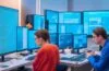 Ein hell erleuchteter moderner Arbeitsplatz mit motiviert arbeitenden Entwicklerinnen und Entwicklern vor mehreren Bildschirmen, die konzentriert und engagiert Rust-Code für sichere Windows-Treiber schreiben, umgeben von warmem Tageslicht, das eine angenehme und freundliche Atmosphäre schafft.