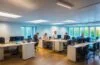 Ein hell erleuchtetes, modernes Büro mit einem freundlichen Team von Mitarbeitenden in angeregter, aber konzentrierter Kommunikation, das gemeinsam an Laptops und Dokumenten arbeitet und dabei Achtsamkeit und Vertrauen ausstrahlt – Sinnbild für gelebte IT-Sicherheit und den Schutz vor Social-Engineering-Angriffen.
