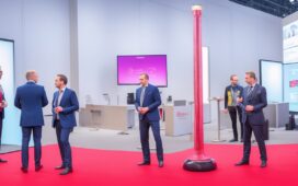 Hell ausgeleuchteter, moderner Messestand mit vereinzelten, nachdenklich wirkenden IT-Profis in Business-Kleidung, die mit leichtem Abstand durch geräumige, stilvoll gestaltete Flure einer Technologie-Konferenz schlendern, während warme Sonneneinstrahlung die Szene freundlich und einladend durchflutet und zugleich eine spürbare Ruhe und Reflexion über den Wandel der Digitalbranche vermittelt.