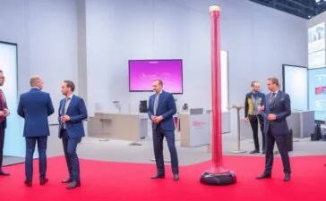 Hell ausgeleuchteter, moderner Messestand mit vereinzelten, nachdenklich wirkenden IT-Profis in Business-Kleidung, die mit leichtem Abstand durch geräumige, stilvoll gestaltete Flure einer Technologie-Konferenz schlendern, während warme Sonneneinstrahlung die Szene freundlich und einladend durchflutet und zugleich eine spürbare Ruhe und Reflexion über den Wandel der Digitalbranche vermittelt.