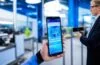 Ein heller, lebendiger Arbeitsbereich mit einem Android-Smartphone in der Hand eines lächelnden Entwicklers, umgeben von natürlichem Tageslicht und modernen technischen Geräten, die eine Atmosphäre von innovation, sicherheit und digitalem Fortschritt vermitteln.