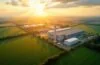 Sonnendurchflutete Landschaft mit modernen Industrierhallen und weitläufigen Freiflächen nahe einer kleinen Gemeinde im Grünen, die durch warme Lichtstrahlen und natürliche Farbnuancen eine hoffnungsvolle Zukunft des digitalen Wandels und nachhaltiger Infrastruktur symbolisieren.