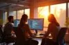 Ein warm beleuchtetes, modernes Entwicklerbüro mit fokussierten Programmiererinnen und Programmierern vor hellen Bildschirmen, die im Sonnenlicht eines großen Fensters Echtzeit-Daten und KI-Modelle visualisieren, und so die lebendige Brücke zwischen künstlicher Intelligenz und Webentwicklung reflektieren.
