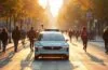 Ein sonnendurchflutetes Stadtbild mit einem eleganten, modernen Robotaxi von Waymo, das sicher und gelassen durch eine belebte, vielfältige Straßenszene mit Fußgängern und Radfahrern gleitet, während warme Lichtreflexe auf den glänzenden Karosserien und lebendige urbane Details eine freundliche, vertrauensvolle Atmosphäre schaffen.