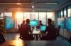 Ein modernes, hell erleuchtetes Büro mit konzentrierten Fachleuten in dynamischer Diskussion vor großen Bildschirmen, die komplexe Low-Code-Plattformen mit warmem Tageslicht durch bodentiefe Fenster umgeben und eine Atmosphäre von Innovation, Zusammenarbeit und zukunftsweisender Sicherheit ausstrahlen.