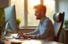Ein strahlend helles Büro mit natürlichem Tageslicht, in dem ein lächelnder UX-Designer konzentriert vor mehreren Bildschirmen sitzt und mit warmen Farben und klar strukturierten Layouts digitale Interfaces gestaltet, die Vertrauen und positive Emotionen ausstrahlen.