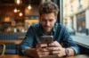 Helle, einladende Aufnahme eines jungen Mannes in urbanem Café-Ambiente, der mit konzentriertem, aber entspanntem Blick sein Android-Smartphone prüft, umgeben von warmem Tageslicht und dezenten technologischen Details, die die vertraute Alltagsnutzung symbolisieren und zugleich die unterschwellige Gefahr digitaler Malware subtil mitschwingen lassen.