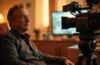 Ein lichtdurchflutetes Filmset-Interieur mit James Cameron in entspannter Pose, umgeben von moderner Kamera- und KI-Technologie, das die harmonische Verbindung von menschlicher Kreativität und innovativen digitalen Werkzeugen in warmen, einladenden Farben einfängt.