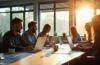 Ein hell erleuchtetes, modernes Büro mit einem entspannten Entwicklerteam, das konzentriert an Laptops arbeitet, während natürliche Sonnenstrahlen durch große Fenster warmes Licht auf saubere Schreibtische mit Code- und Workflow-Diagrammen werfen und so eine freundliche, produktive Atmosphäre der Vernetzung und Kostenoptimierung in der Softwareentwicklung vermittelt.