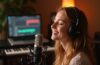 Ein natürlich hell ausgeleuchtetes, warmes Studiofoto einer jungen Musikerin, die mit geschlossenen Augen und sanftem Lächeln entspannt in ein hochwertiges Mikrofon singt, während im Hintergrund verschwommen moderne Musikgeräte und ein Laptop mit Digital Audio Workstation zu erkennen sind – die harmonische Verbindung von menschlicher Kreativität und moderner Technik spürbar in jedem Detail.