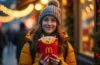 Ein strahlend helles, natürlich beleuchtetes Porträt einer nachdenklichen jungen Frau in urbaner Weihnachtskulisse, die mit warmem Lächeln einen McDonald’s-Essensbeutel in den Händen hält, während sanftes Tageslicht menschliche Nähe und die subtilen Spannungen zwischen Technik und Authentizität einfängt.